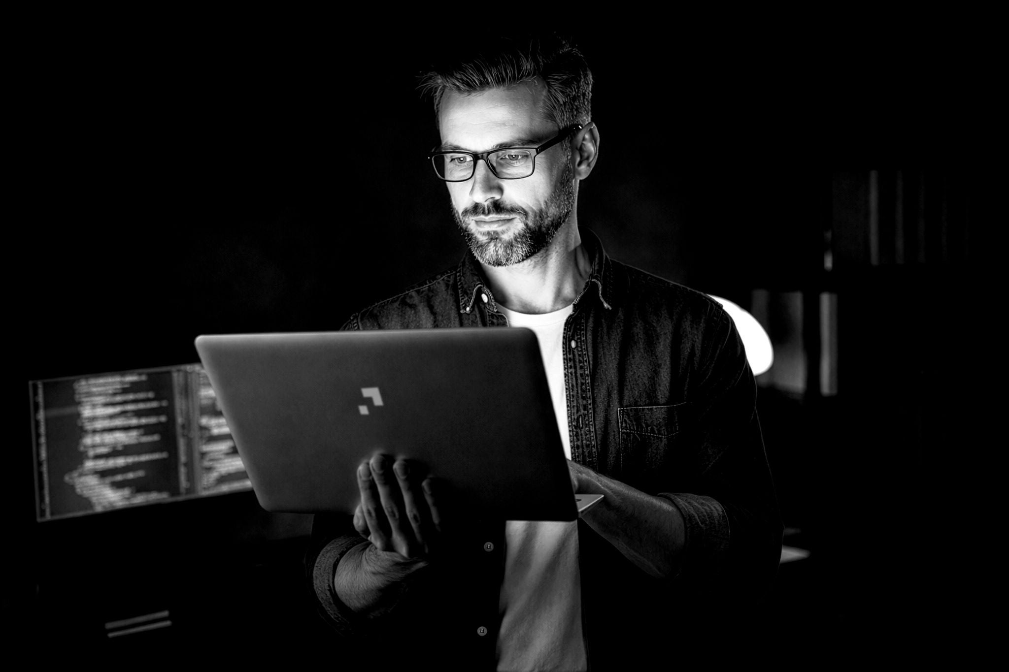 Personne utilisant un ordinateur portable dans un environnement sombre, avec des écrans affichant du code en arrière-plan, illustrant un contexte de travail numérique et technologique